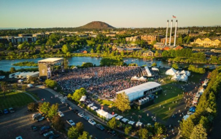Aerial view of Hayden Homes Amphitheater during a concert at sunset, with the Old Mill District, smokestacks, and Pilot Butte visible in the background.