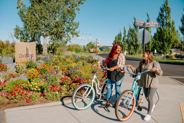 Two girls walk their cruiser bikes side by side on a sidewalk with the Old Mill District sign and iconic smokestacks in the background.